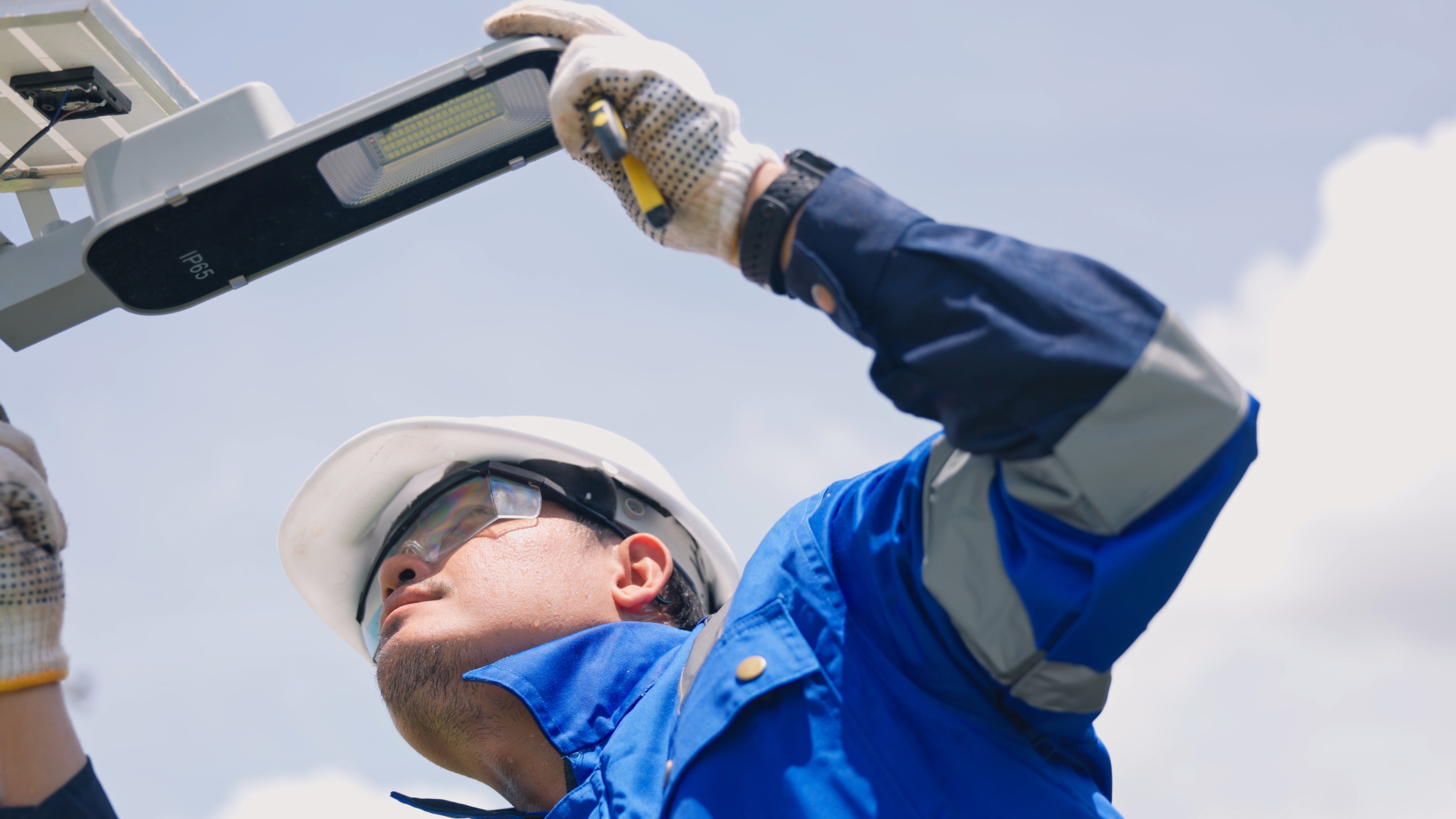 Asian male technician in safety helmet installing or repairing outdoor LED solar street light on pole. Professional industrial electrical maintenance and infrastructure service.